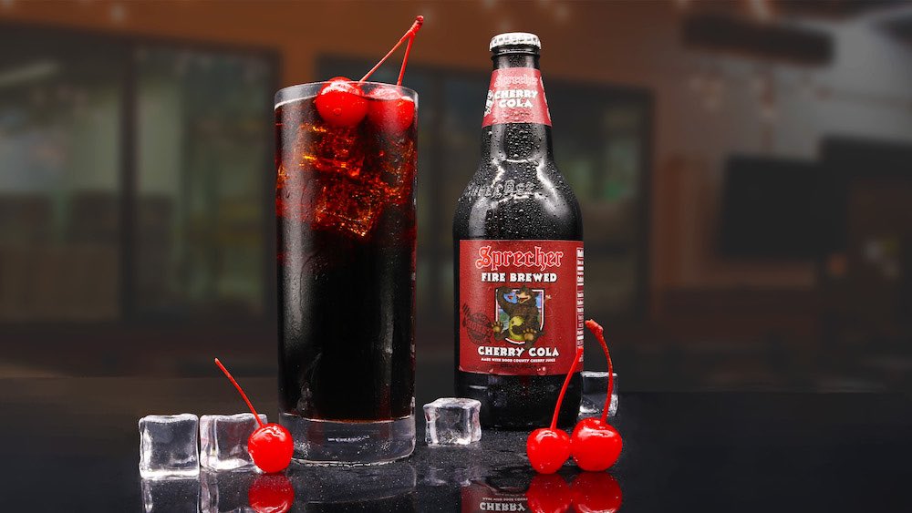 A glass of iced Sprecher Cherry cola with maraschino cherries floating in the cola and in the foreground, and a bottle of Sprecher Cherry Cola to the right