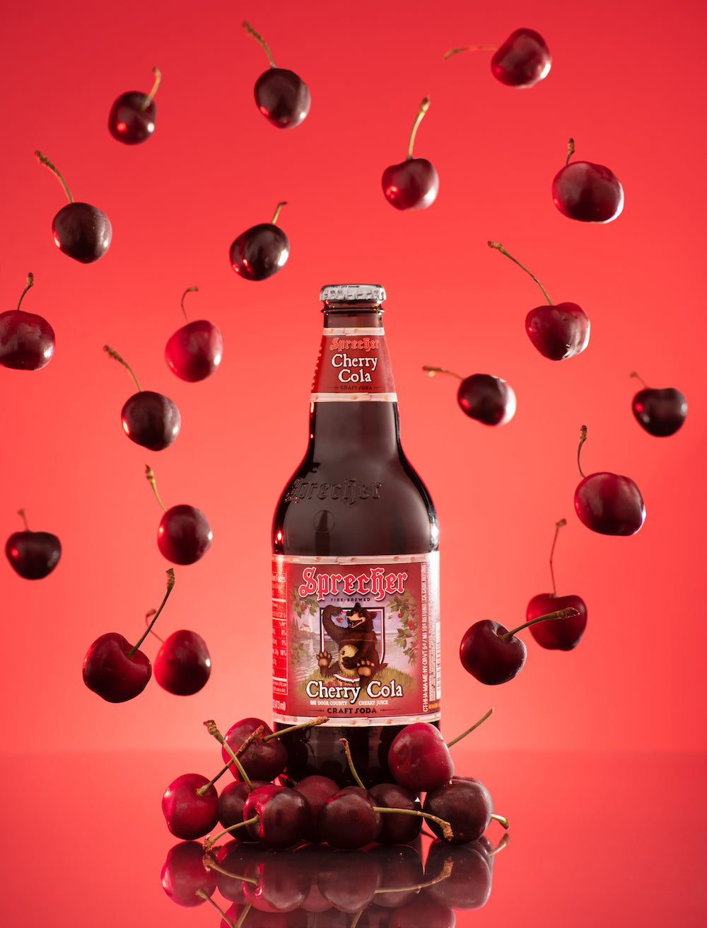 A 16oz bottle of Cherry Cola surrounded by falling cherries with a red background