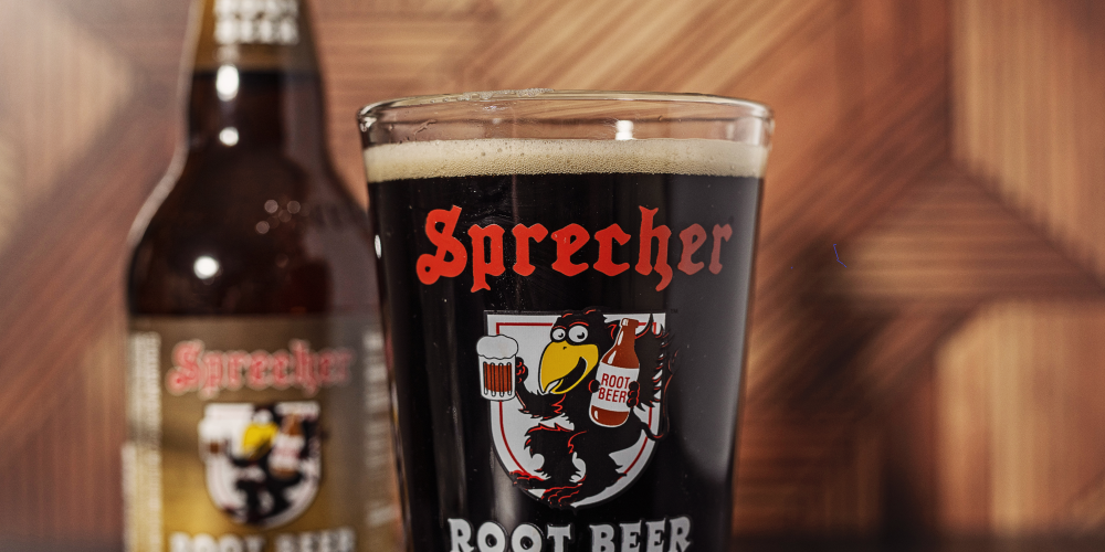 A pint glass full of Sprecher root beer with a 16oz bottle of root beer in the background