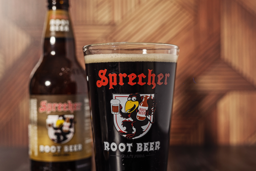 A pint glass full of Sprecher root beer with a 16oz bottle of root beer in the background