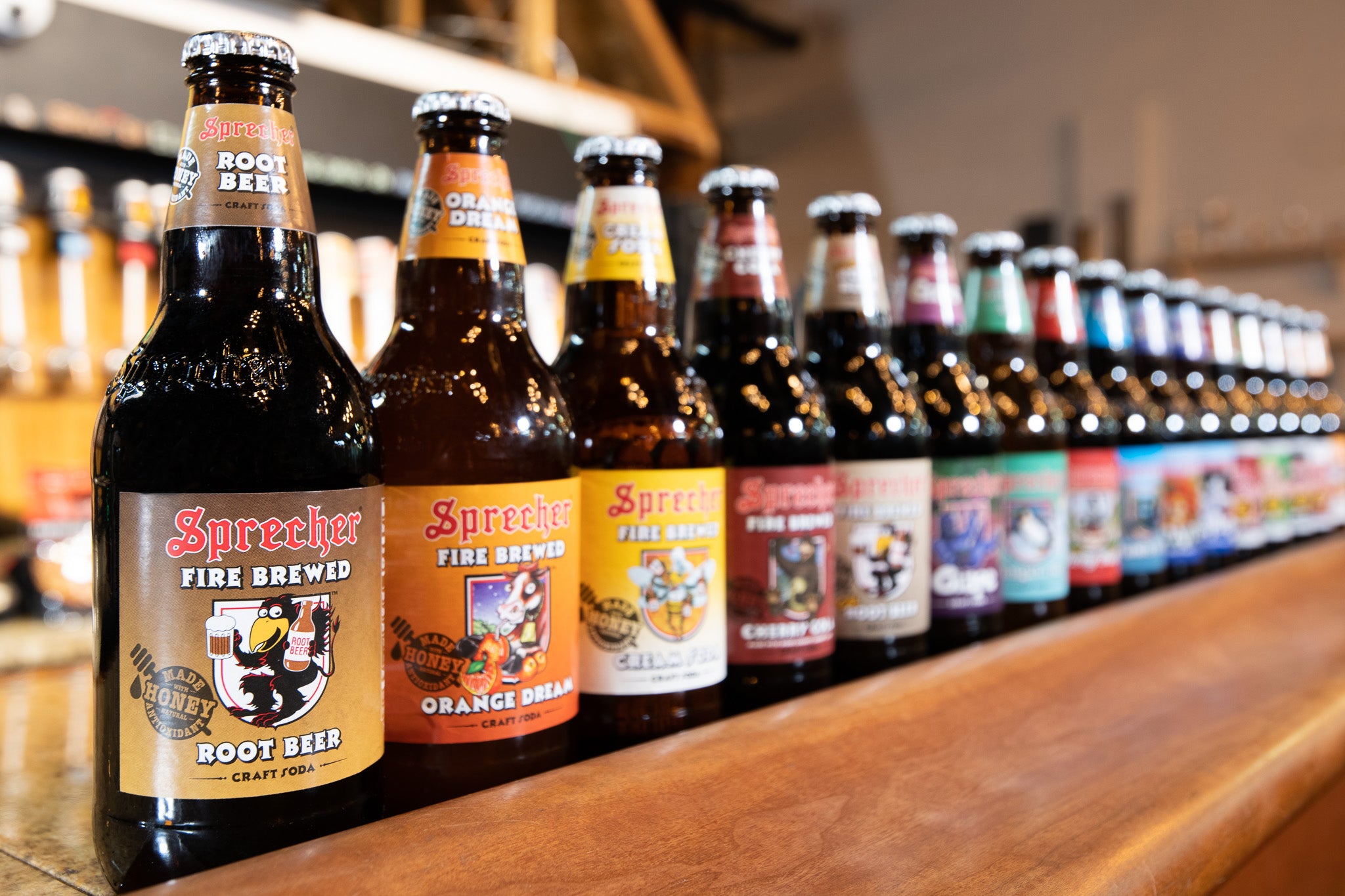 Sprehcer Soda Bottles lined up from left to right: Root Beer, Orange Dream, Cream Soda, Cherry Cola, Low-Cal Root Beer, Grape, Ginger Ale--additional bottles blurred into the background