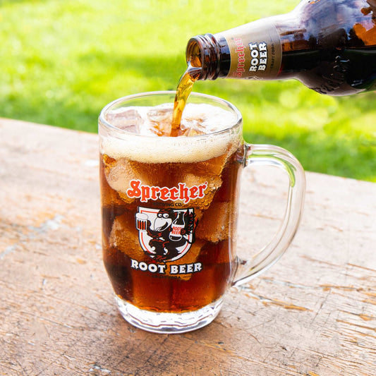 Sprecher Branded Glass Root Beer Mug being filled with Sprecher Root Beer on a picnic table with a grassy green background