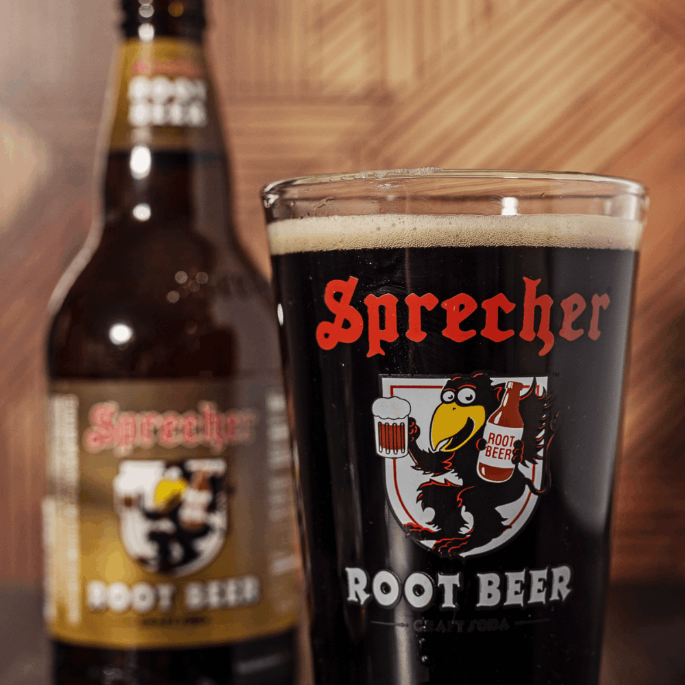 A pint glass of Sprecher Root Beer in the foreground with a blurry bottle of Sprecher Root Beer in the background