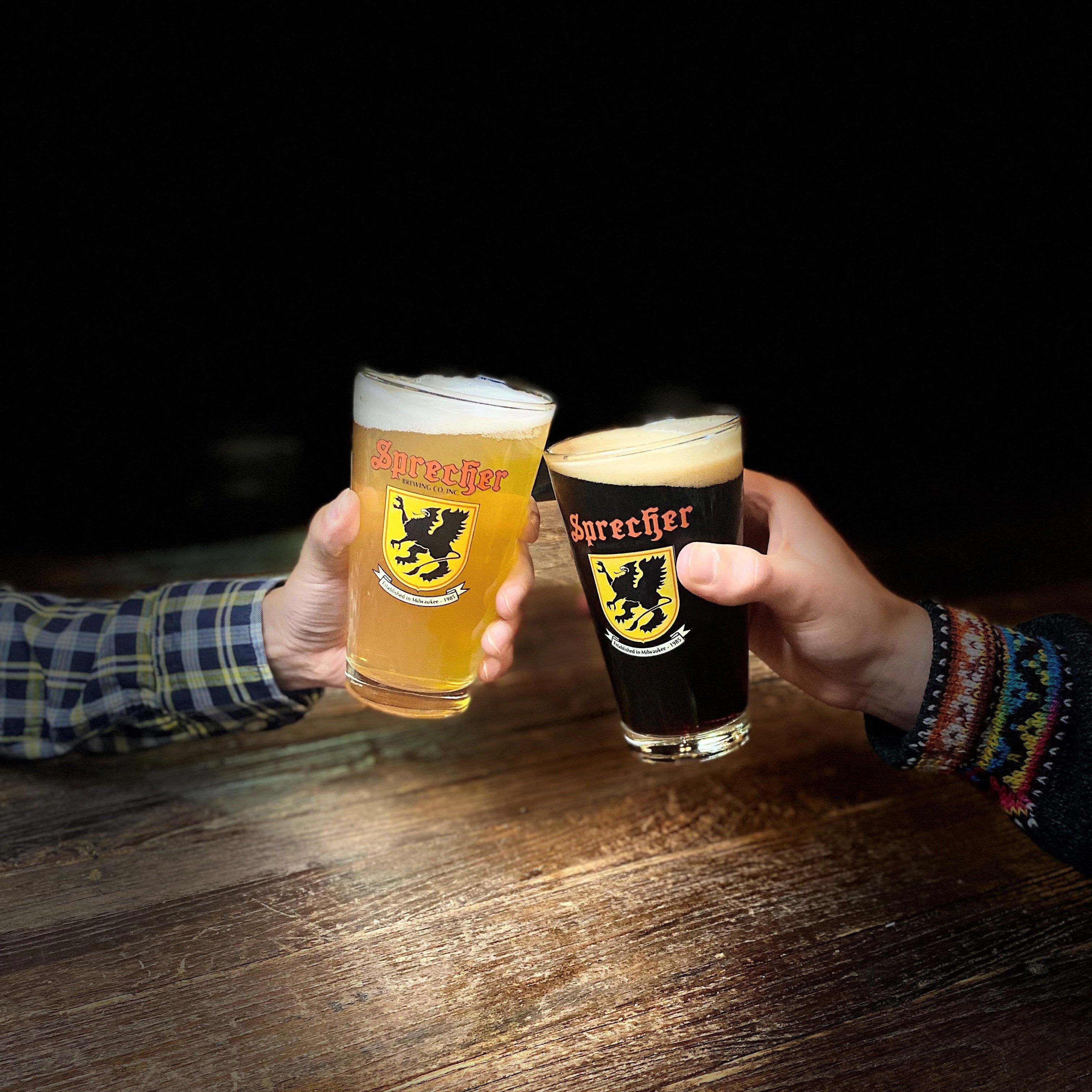 Two hands holding pint glasses with beer at a wooden table. One glass contains a golden beer with a white foamy head and a yellow sprecher logo, and the other contains a dark black beer with a white foamy head and a yellow sprecher logo.