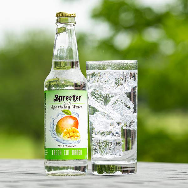 12oz bottle of Fresh Cut Mango Sparkling Water next to a glass of Sparkling Water with an outdoor background