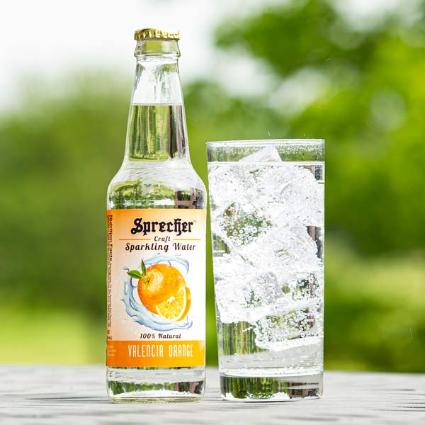 12oz bottle of Sprecher Valencia Orange Sparkling water next to a glass of sparkling water with an outdoor background